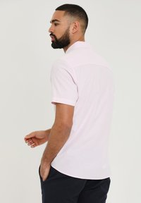Korte mouw polo shirt in lichtroze stof, met een kraag, een gladde textuur en een rechte zoom. Het model heeft zijn handen in de zakken.