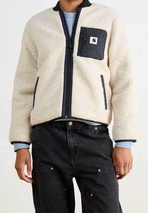 Osoba ubrana w kremową kurtkę polarową z czarnymi wykończeniami i zapinaną na zamek kieszenią na piersi, w połączeniu z czarnymi dżinsami i widocznymi mankietami niebieskiej, długorękawowej koszuli.