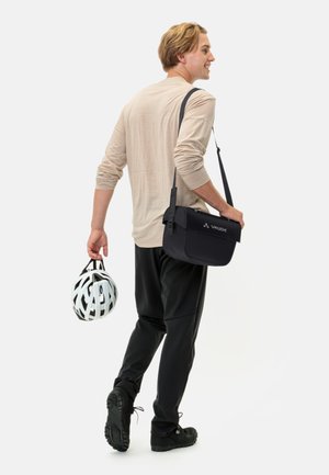 Sac à messager Vaude noir avec sangle réglable, texture lisse et fermeture à rabat, tenu par une personne en tenue décontractée, portant un casque blanc.