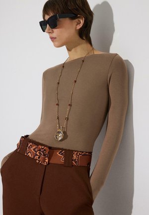 Persona con gafas de sol rectangulares negras, camiseta de manga larga beige, cinturón marrón con estampado, pantalones marrones y collar largo de oro con colgante.