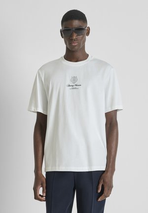 Hombre con gafas de sol negras, camiseta blanca de manga corta con un pequeño logo y pantalones oscuros sobre un fondo claro y liso.