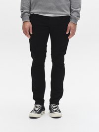 Crne uske hlače glatke teksture, u kombinaciji s crno-bijelim tenisicama s prugama i vezicama, nošene ispod sive veste.