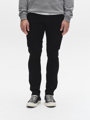 Črne slim-fit hlače z gladko teksturo, obute v črno-bele superge s črtami in vezalkami, nošene pod sivim puloverjem.