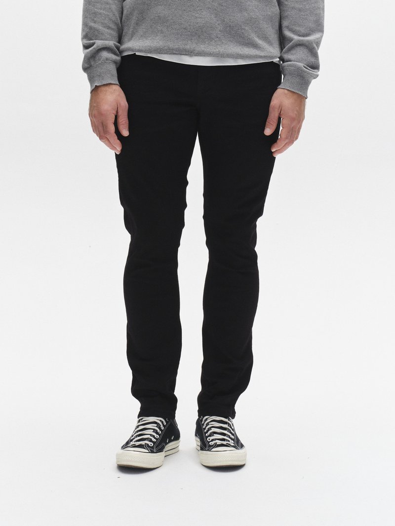 Crne uske hlače glatke teksture, u kombinaciji s crno-bijelim tenisicama s prugama i vezicama, nošene ispod sive veste.