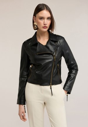 Kvinde iført sort lædermotorcykeljakke med guldlynlås og cremefarvede bukser, poserer foran en neutral baggrund.