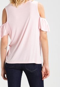 Jasnoróżowa bluzka typu cold-shoulder z falbaniastymi rękawami, miękki materiał, luźny krój i lekko zaokrąglony dół; zestawiona z ciemnoniebieskimi dżinsami.