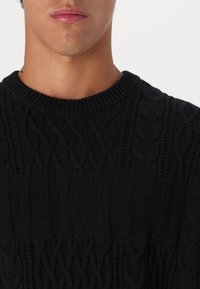 Black knitted sweater featuring a chunky cable design, ribbed neckline, and textured pattern, with prominent brand lettering across the chest.