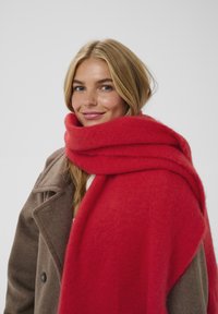 Red wool scarf draped around shoulders, featuring a soft, textured finish. Worn with a light brown coat showcasing visible buttons.