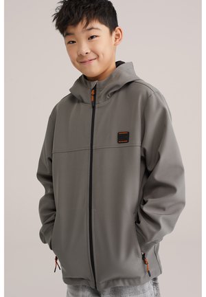 Mladi dječak s rukama u džepovima nosi sivu kapuljaču na patentni zatvarač s narančastim zatvaračima i malim patchom na prsima.