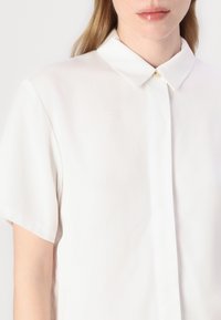 Camicia bianca a maniche corte realizzata in tessuto morbido, con colletto a punta e chiusura con bottone al collo.