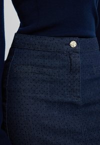 Laivastonsininen farkkuhame, jossa on teksturoitu pilkkukuvio, kiiltävä hopeanvärinen nappi ja kaksi etutaskua.