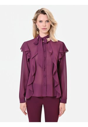 Donna che indossa una camicetta viola trasparente con volant e collo annodato, abbinata a pantaloni viola coordinati su uno sfondo bianco semplice.