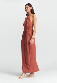 Rudos spalvos, be rankovių halter tipo suknelė su draped priekiu, surištu liemens detalė ir ilgu, laisvai krentančiu sijonu. Pora su lengvais atvirais bateliais.