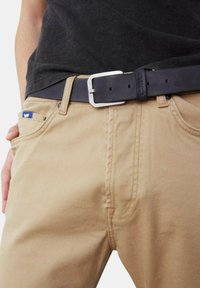 Pantaloni di cotone beige con tasche frontali, presentano una cintura nera con una fibbia in argento e un piccolo logo blu sulla tasca sinistra.