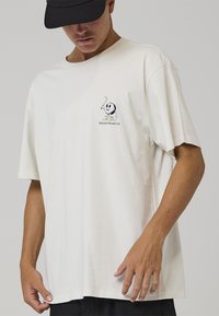 Kremfarvet, løstsiddende t-shirt lavet af blød bomuld med en lille broderet figur på venstre bryst.