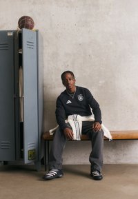 Mladic u sportskoj odjeći sjedi na klupi pored otvorenog ormarića s loptom u mreži na vrhu, protiv pozadine od betonskog zida.