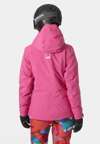 Fuchsijak s kapucí, texturovaný povrch a logo na zadní straně; v kombinaci s mnohobarevnými vzorovanými lyžařskými kalhotami.