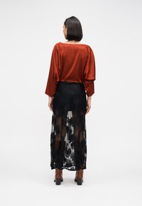 Μπλούζα από μετάξι, σε σκουριά χρώμα, με φαρδιά μανίκια, συνδυασμένη με διαφανή φούστα από μαύρη δαντέλα με λουλουδένια σχέδια και καφέ μπότες με τετράγωνο τακούνι.