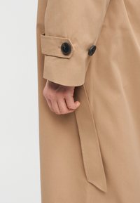 Manche d'un trench-coat beige avec des boutons noirs et une sangle de ceinture, montrant une main reposant à côté du manteau.