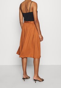 Woman wearing a black sleeveless top, rust-colored midi skirt with belt, and leopard print heeled mules, standing against a plain background.