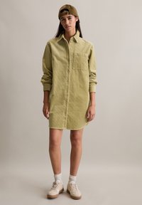 Lichtgroene corduroy jurk met knoopsluiting aan de voorkant, lange mouwen, borstzak, gedragen met een bruine pet en beige sneakers.