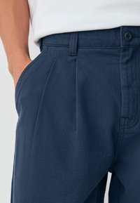 Navy blue trousers made of textured fabric, featuring pleats, a button closure, and side pockets. Close-up view of the waistband area.