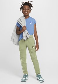 Maglietta Nike blu con logo bianco, felpa grigia appoggiata sulla spalla, pantaloni cargo verdi con tasche e sneakers verdi.