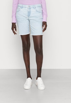 Personne portant un short en jean bleu clair avec des ourlets bruts, un haut manches longues lavande et des baskets blanches, debout sur un sol gris.
