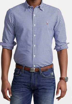 Mand iført blå og hvid ternet skjorte med knapper, opbukket ærmer, brun læderbælte, blå jeans og armbåndsur på venstre håndled.