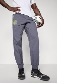 Mand wearing grå Manchester City track pants og sorte sportssko, holder en sort og hvid fodbold mod en ensfarvet baggrund.