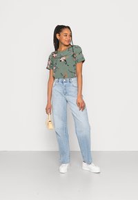 Top de manga curta com estampado floral verde, jeans de perna reta azul claro, sapatilhas brancas de plataforma e uma pequena bolsa amarela.