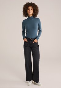 Pull à col roulé en teal, à manches longues, au design ajusté, associé à un jean noir à jambes larges, rehaussé d'une ceinture noire et de baskets claires.