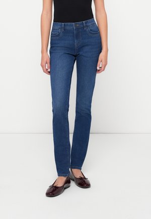 Femme portant un jean bleu ajusté et des ballerines rouge foncé, debout devant un fond blanc.