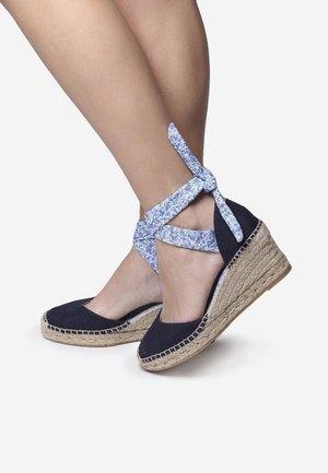 Navy canvas espadrille wedges dengan platform jute alami dan tali pergelangan kaki berbunga biru. Memiliki bagian depan tertutup dan detail anyaman.