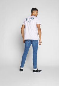 Camiseta rosa clara con un gráfico de "Billabong" en la parte trasera, combinada con jeans ajustados azules y zapatillas negras. La textura parece suave.