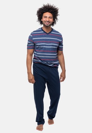 Ein Mann mit lockigem Haar lächelt, trägt ein blau-rot gestreiftes Kurzarmshirt und marineblaue Hose, geht barfuß auf weißem Hintergrund.