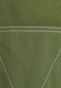 Nära bild av olivgrön tyg med vita dubbelsömmar över det strukturerade materialet.