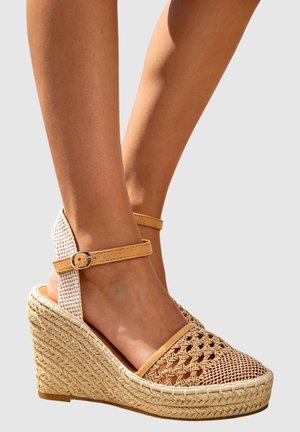 Sandales compensées tressées couleur fauve avec brides à la cheville et plateforme en corde tressée, portées pieds nus sur un fond neutre.