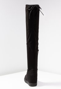 Botte noire en daim montant jusqu'au genou avec une texture lisse, bout arrondi, semelle plate et une fermeture à nouer en haut pour un ajustement sécurisé.