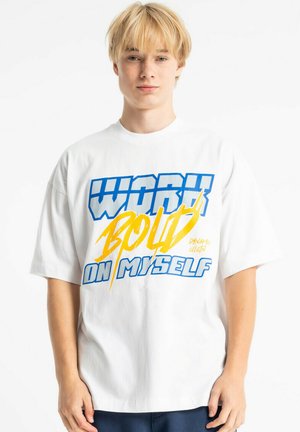 Młoda osoba o blond włosach, ubrana w białą koszulę z niebiesko-żółtym napisem „Work Bold on Myself” na jednolitym tle.