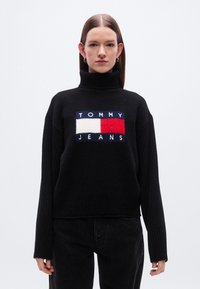 Pull à col roulé noir avec une texture côtelée, orné au centre d'un logo brodé en rouge, blanc et bleu indiquant "TOMMY JEANS".