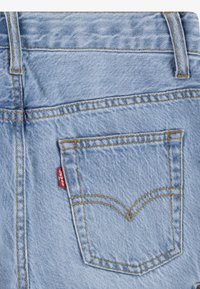 Poche arrière d’un jean en denim bleu clair avec des surpiqûres jaunes et une étiquette rouge Levi’s visible sur la couture de la poche.