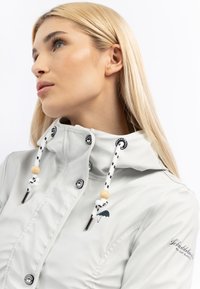 Jachetă impermeabilă de culoare gri deschis, cu guler înalt și șnururi de contrast negre și albe. Are mărgele din lemn și un logo brodat cu umbrele.