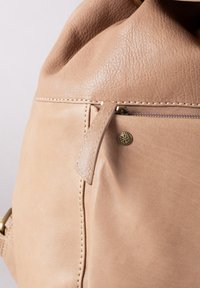 Beige leather handbag featuring a zippered pocket, visible stitching, and a decorative metallic accent. Soft texture with a slouchy design.