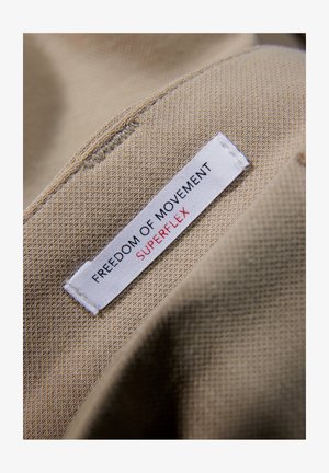 Beige kangas, jolla on tekstuuripinta, jossa on valkoinen etiketti, jossa lukee mustalla ja punaisella fontilla "VAPAASTI LIIKKUMINEN SUPERFLEX".