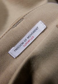 Close-up van beige stof met een wit kledinglabel waarop "FREEDOM OF MOVEMENT SUPERFLEX" staat, vastgenaaid in de naad.