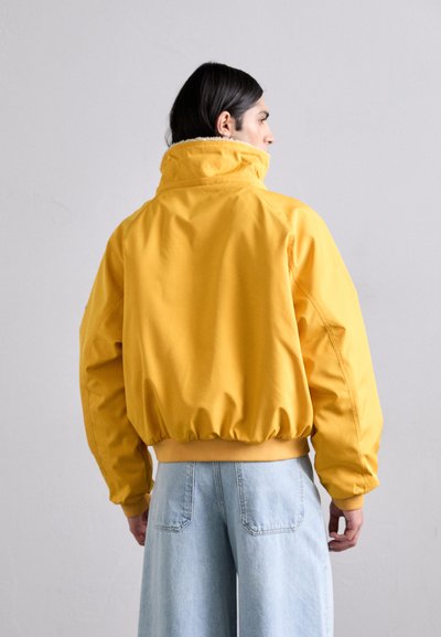 Yellow, padded outerwear jacket with a high collar and elastic hem. Features a smooth texture and casual fit, paired with light blue jeans.