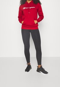 Červená mikina s kapucí a bílým logem "Champion", s klokankou, ve spojení s černými legínami a černými sportovními botami.