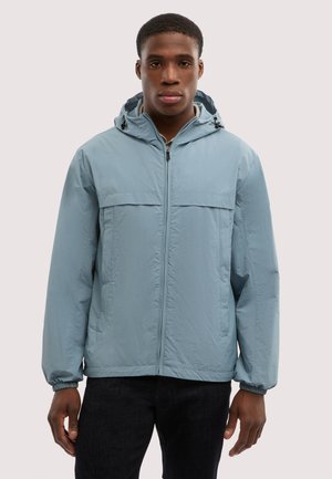 Junger Mann trägt eine hellblaue Kapuzenjacke mit Reißverschluss und schwarze Hose, steht vor einem einfarbigen hellen Hintergrund.