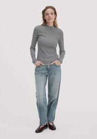 Grauer Langarmshirt mit hohem Kragen, kombiniert mit hellblauen, weiten Jeans und burgunderfarbenen Slippern, Hände in den Taschen. Einfarbiger Hintergrund.